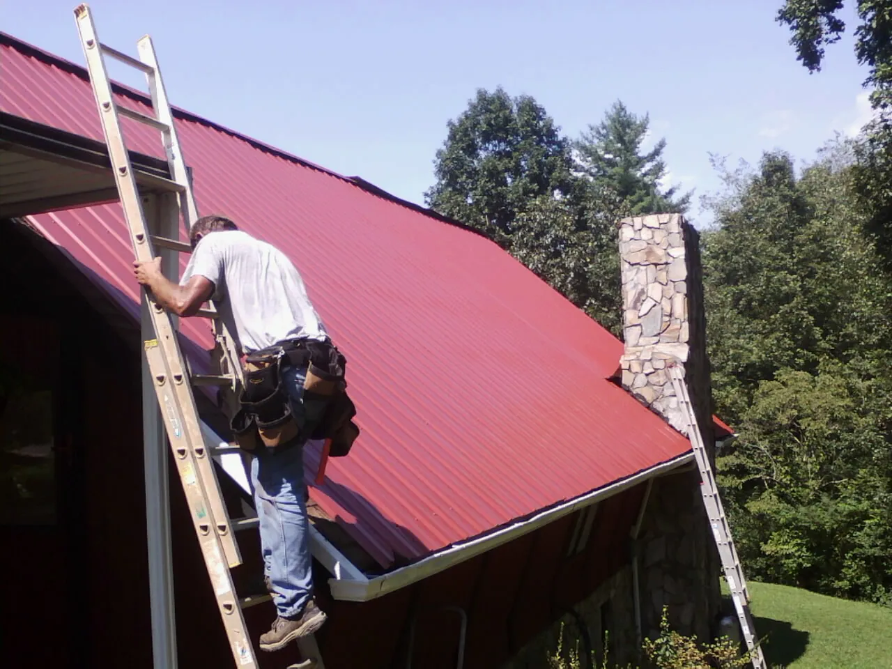Roofer on a ladder beside a red metal roof and stone chimney — Soffit & Fascia Repair by Roofing Brothers in Mankato